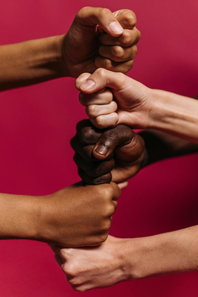 Three hands of different skin tones clasped together in unity against a pink background, symbolizing community and support.